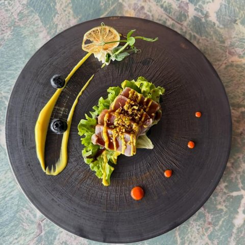 Tataki di tonno scottato con granella di pistacchio, salsa teriyaki e maionese al mango, su foglia di lattuga, visto dall'alto su piatto nero